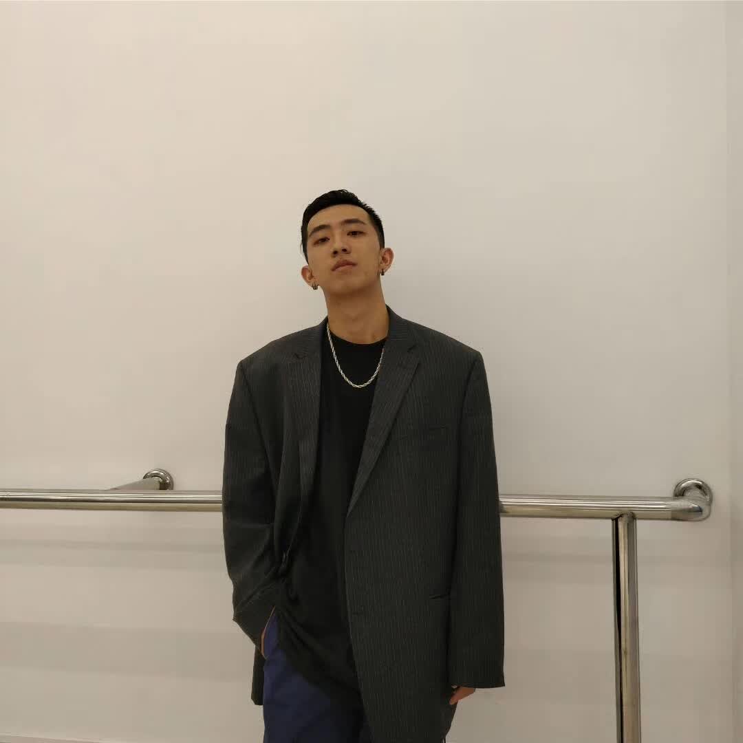 Posing in a dark blazer and silver necklace against a light wall.