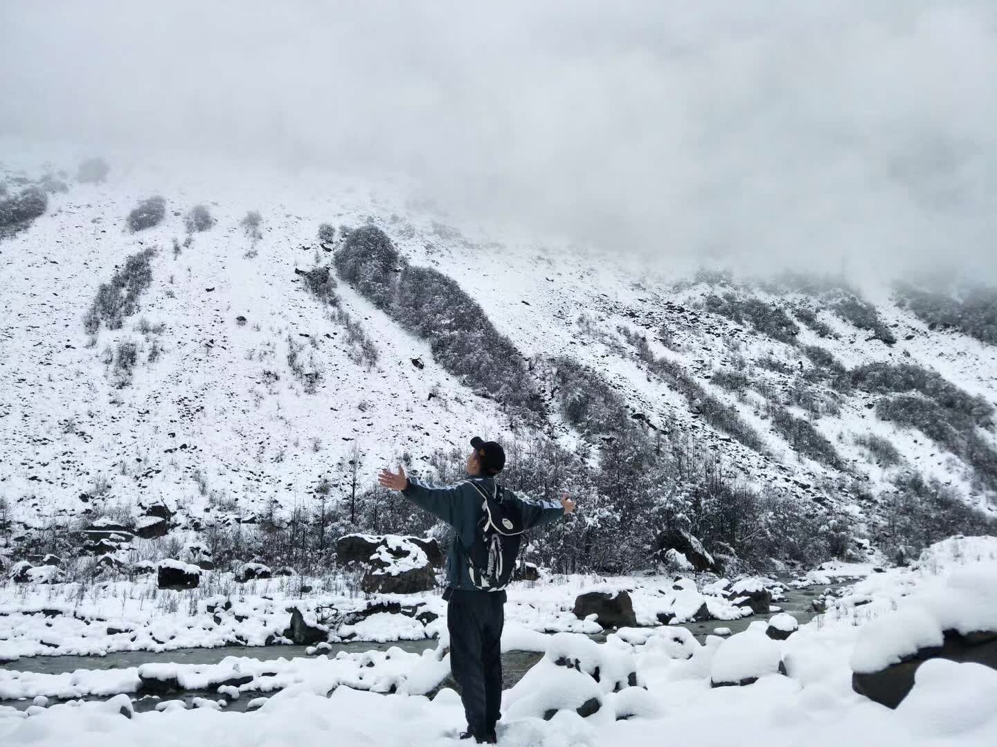 A person with outstretched arms beside a stream in a snow-covered mountain landscape.