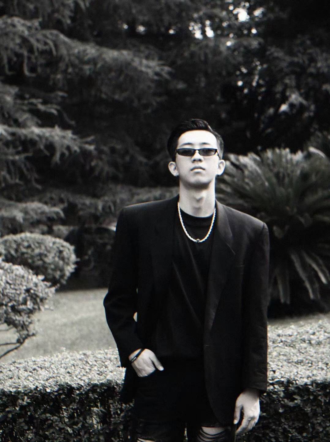 A black-and-white outdoor portrait wearing sunglasses, a dark blazer, and a chain necklace.