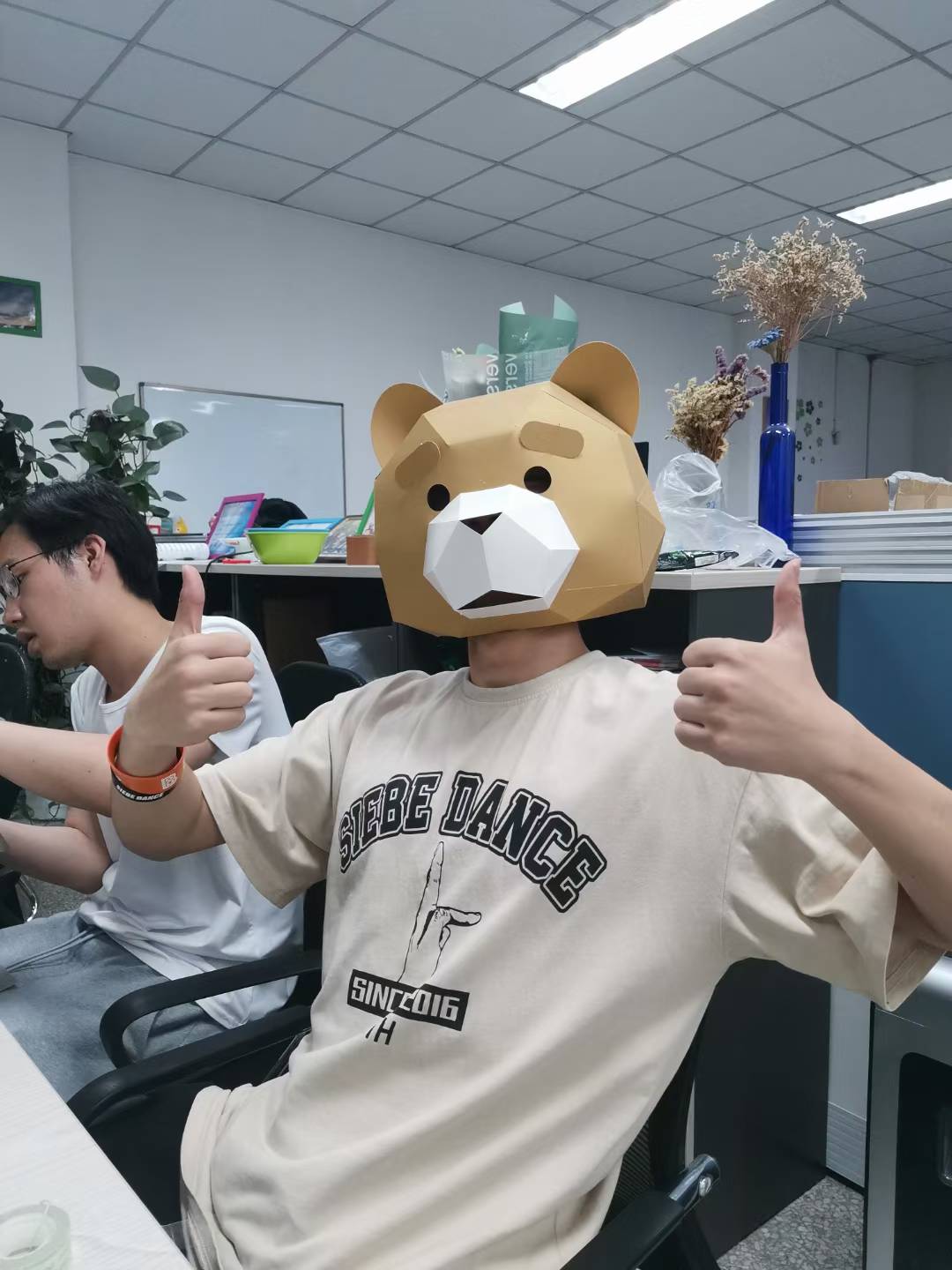 A person wearing a geometric bear mask and giving two thumbs up in a lab office.