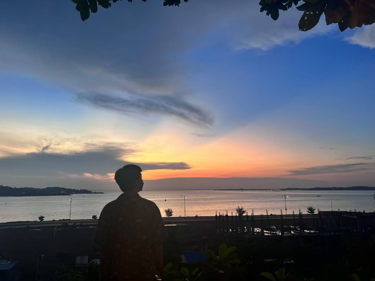 A silhouette looking out over the water beneath a colorful sunset sky.