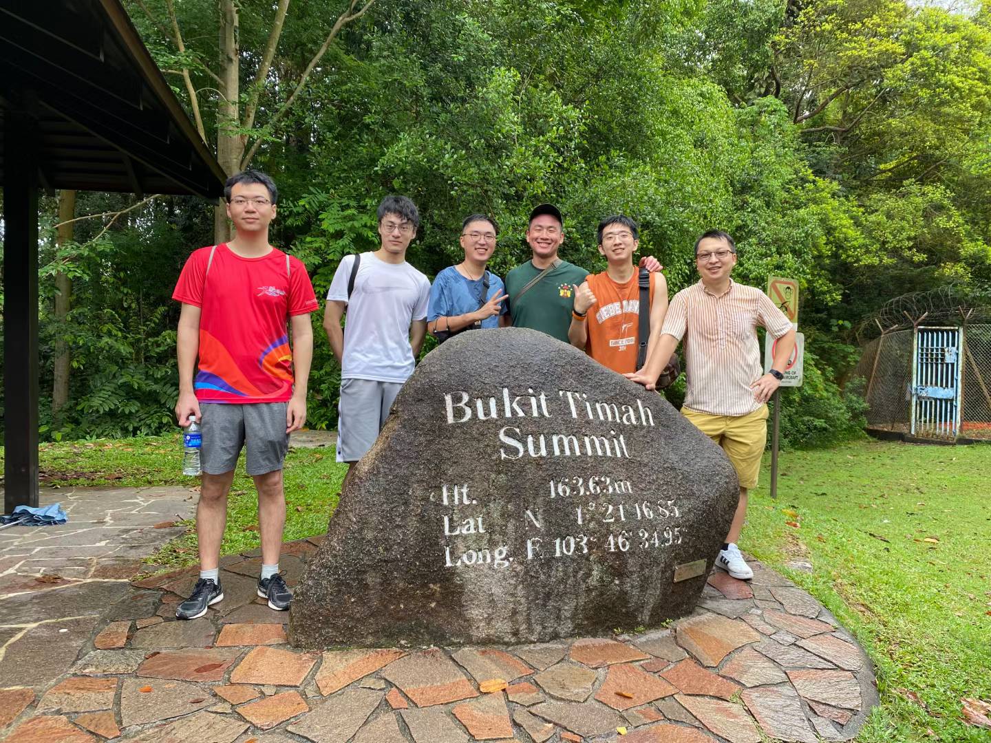 Six hikers posing behind the Bukit Timah Summit stone marker surrounded by trees.