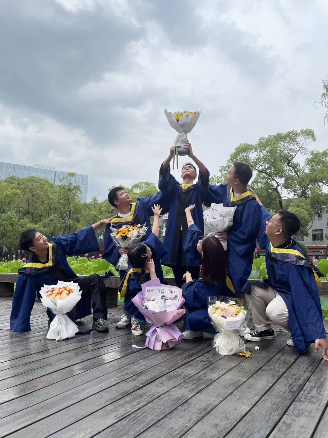Graduates in blue gowns posing with bouquets on a wooden deck, with one bouquet held overhead.