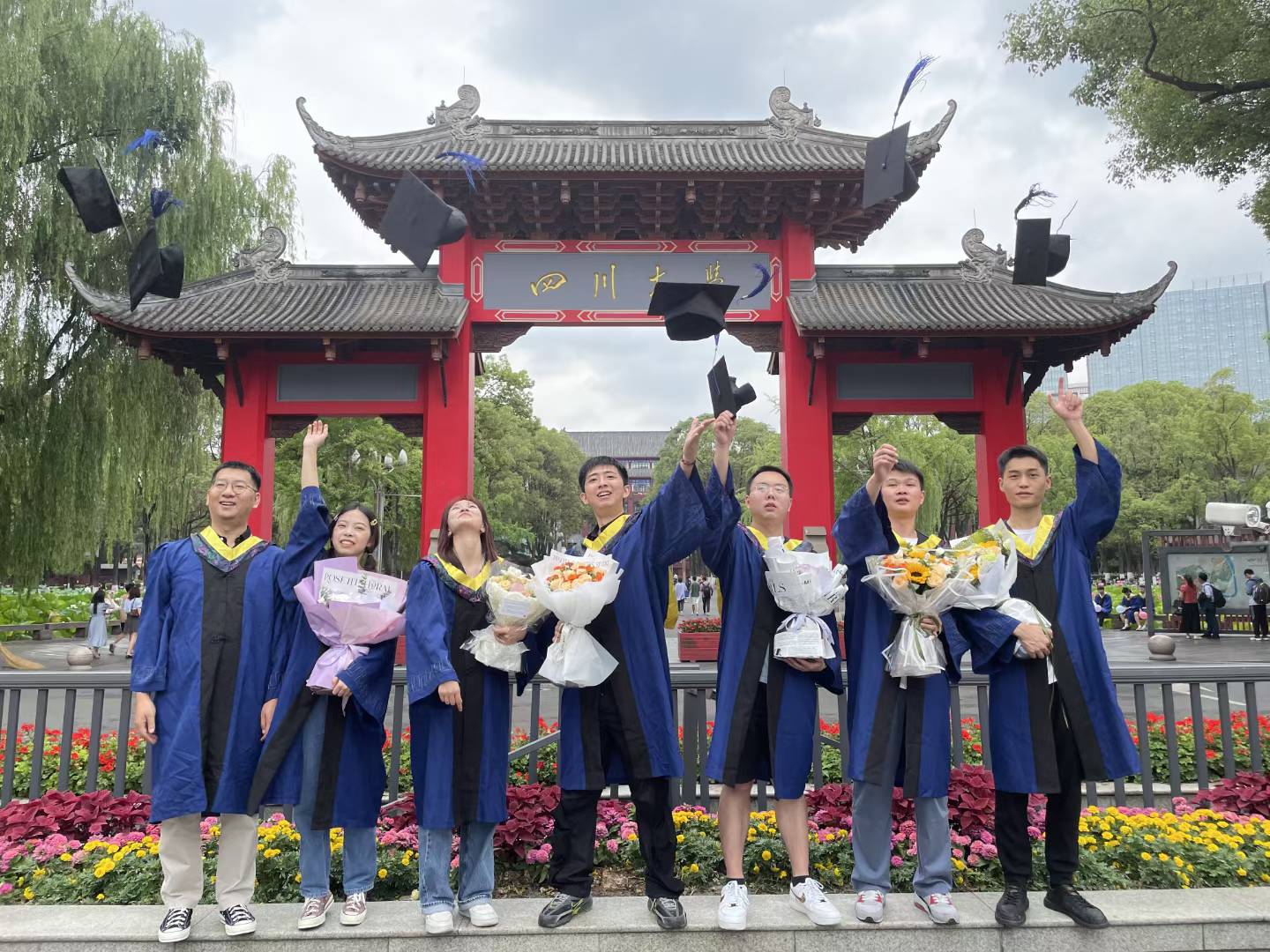 Graduates in blue gowns tossing their caps in front of the Sichuan University gate.