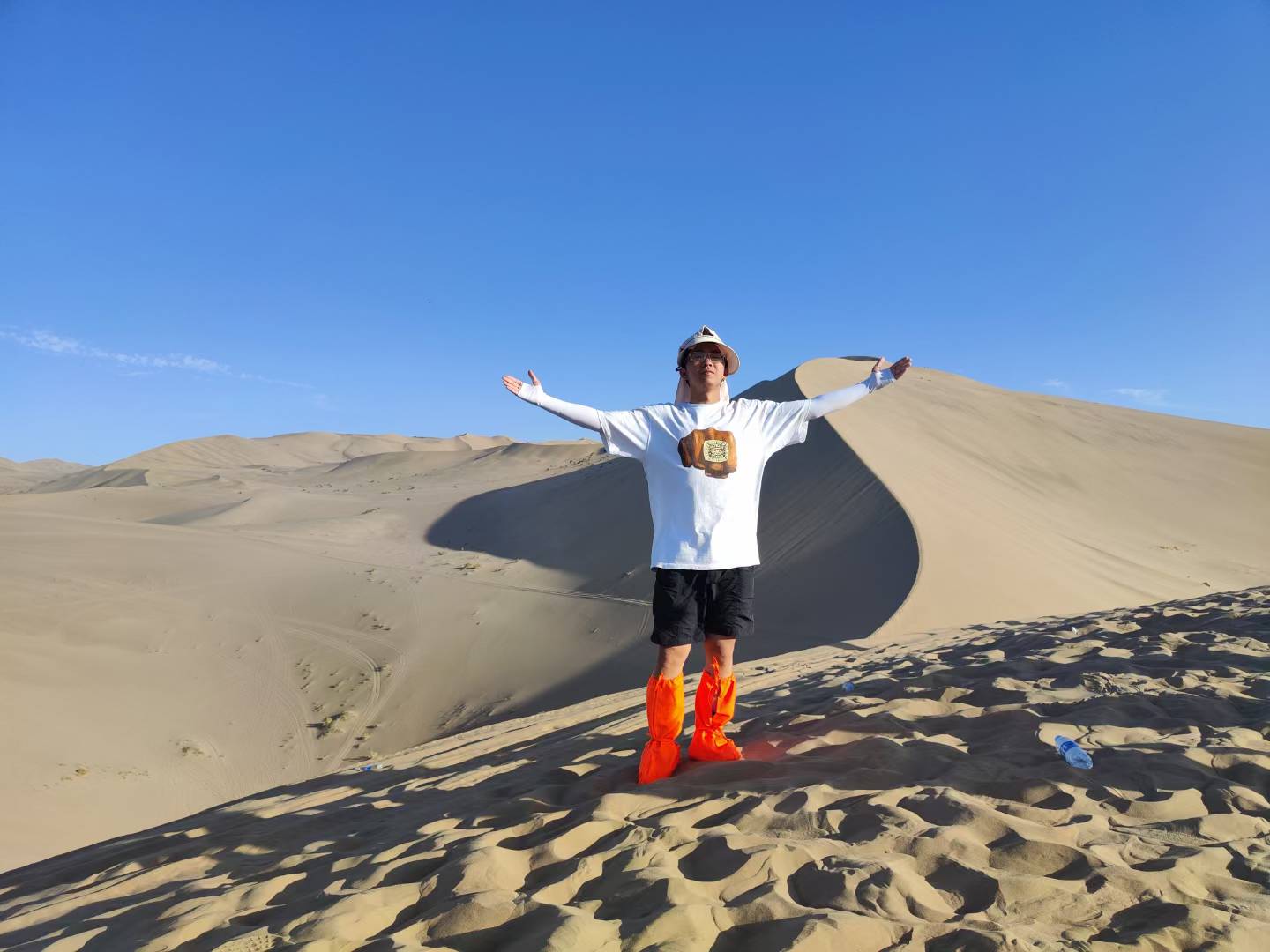 A person standing with outstretched arms on sand dunes beneath a clear blue sky.