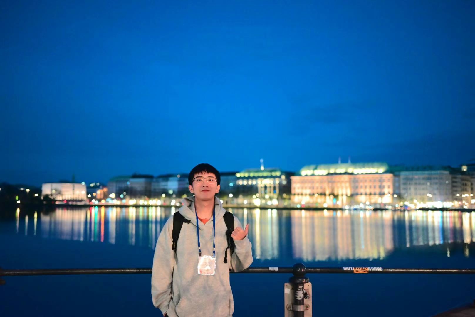 A person wearing a conference badge beside the waterfront at dusk, with city lights reflected in the water.