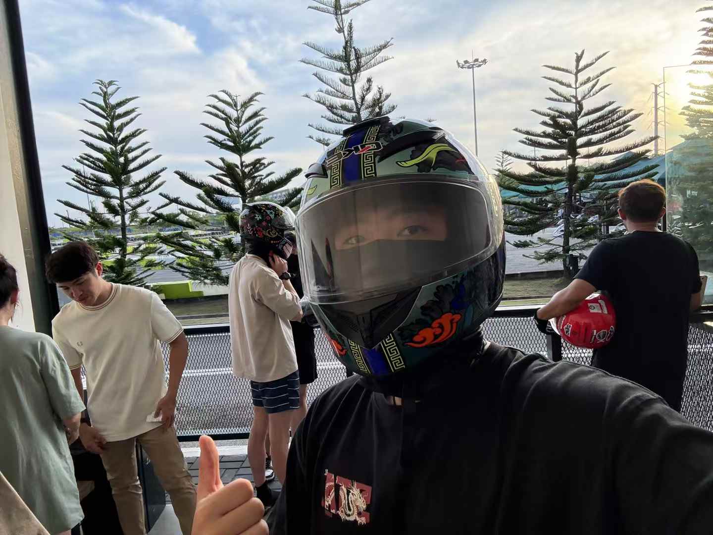A selfie wearing a colorful racing helmet and giving a thumbs up beside a go-kart track.