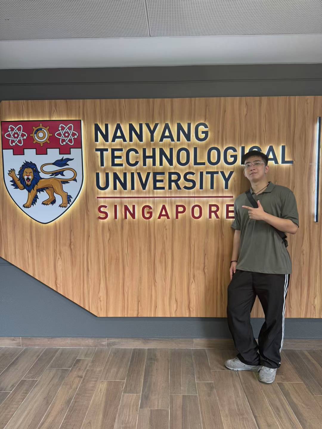 A person posing beside the Nanyang Technological University sign and crest.