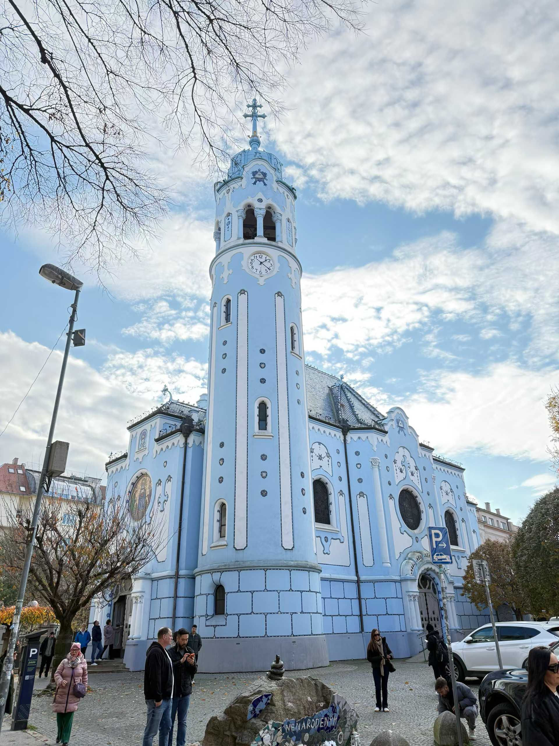 A pale blue church with a tall clock tower beneath a partly cloudy sky.