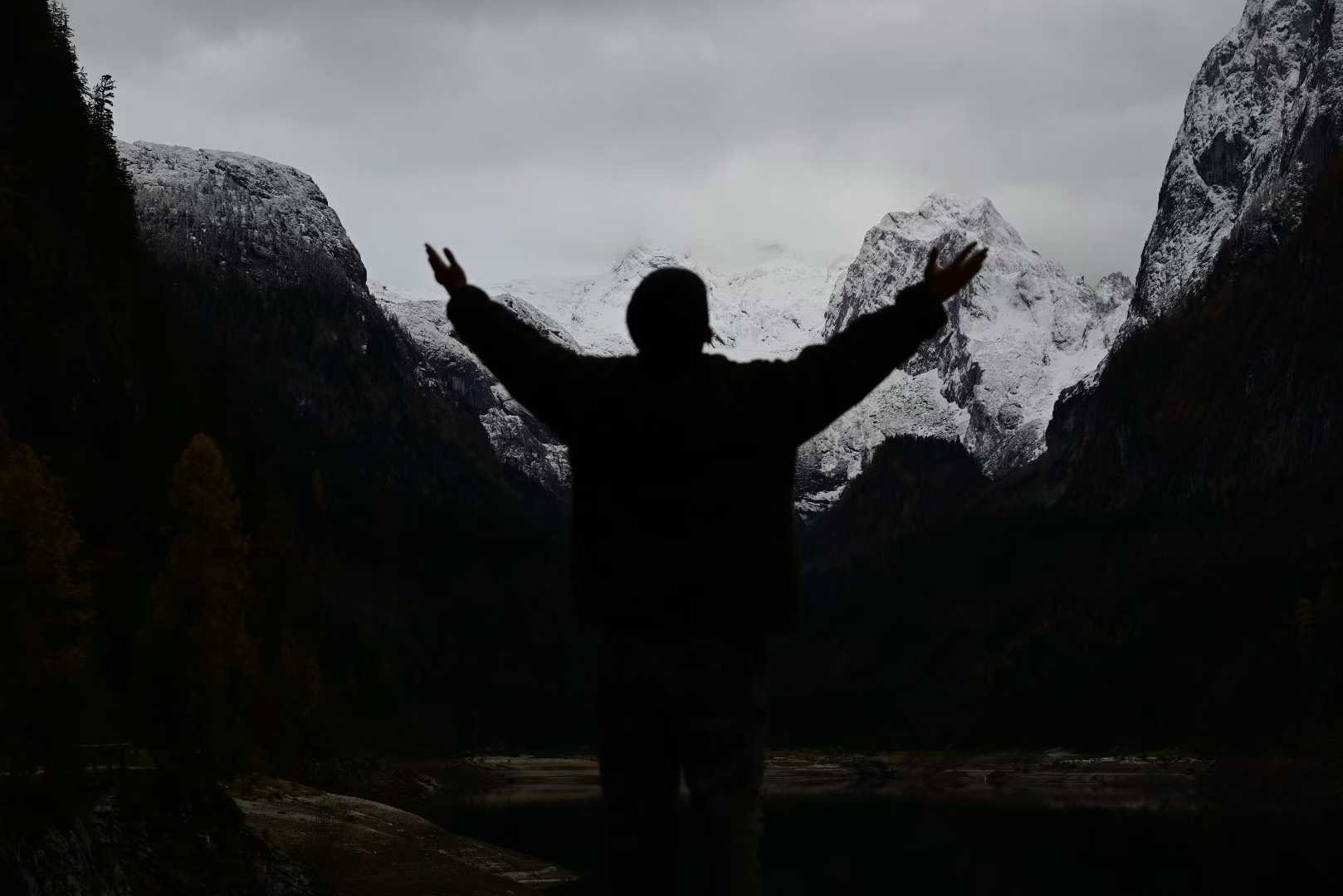 A silhouette with outstretched arms beside a lake surrounded by snow-covered mountains.