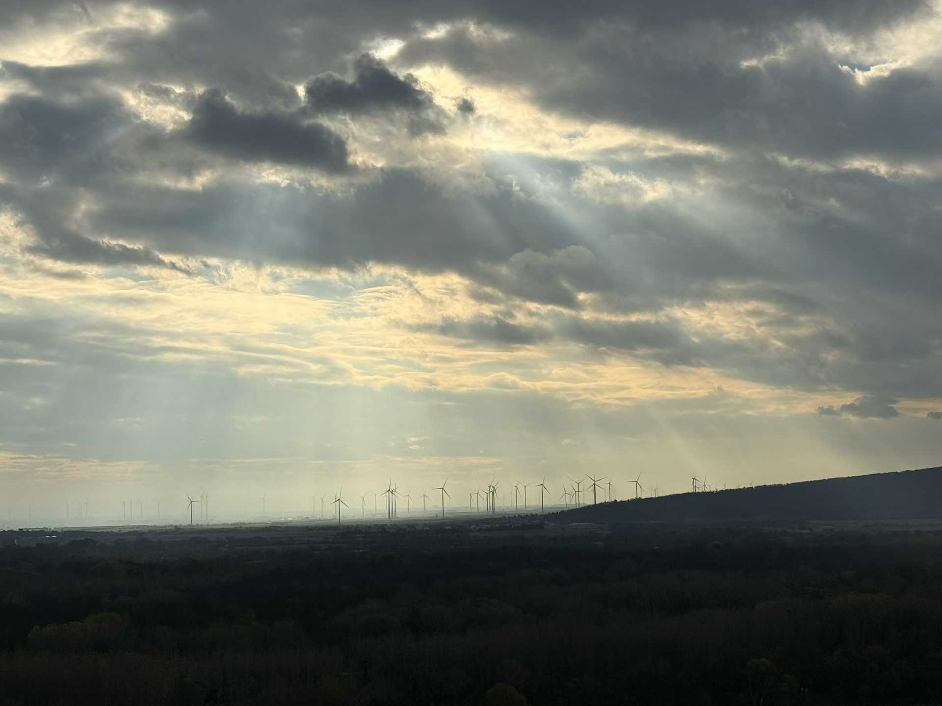 Sunbeams breaking through clouds above a landscape dotted with wind turbines.