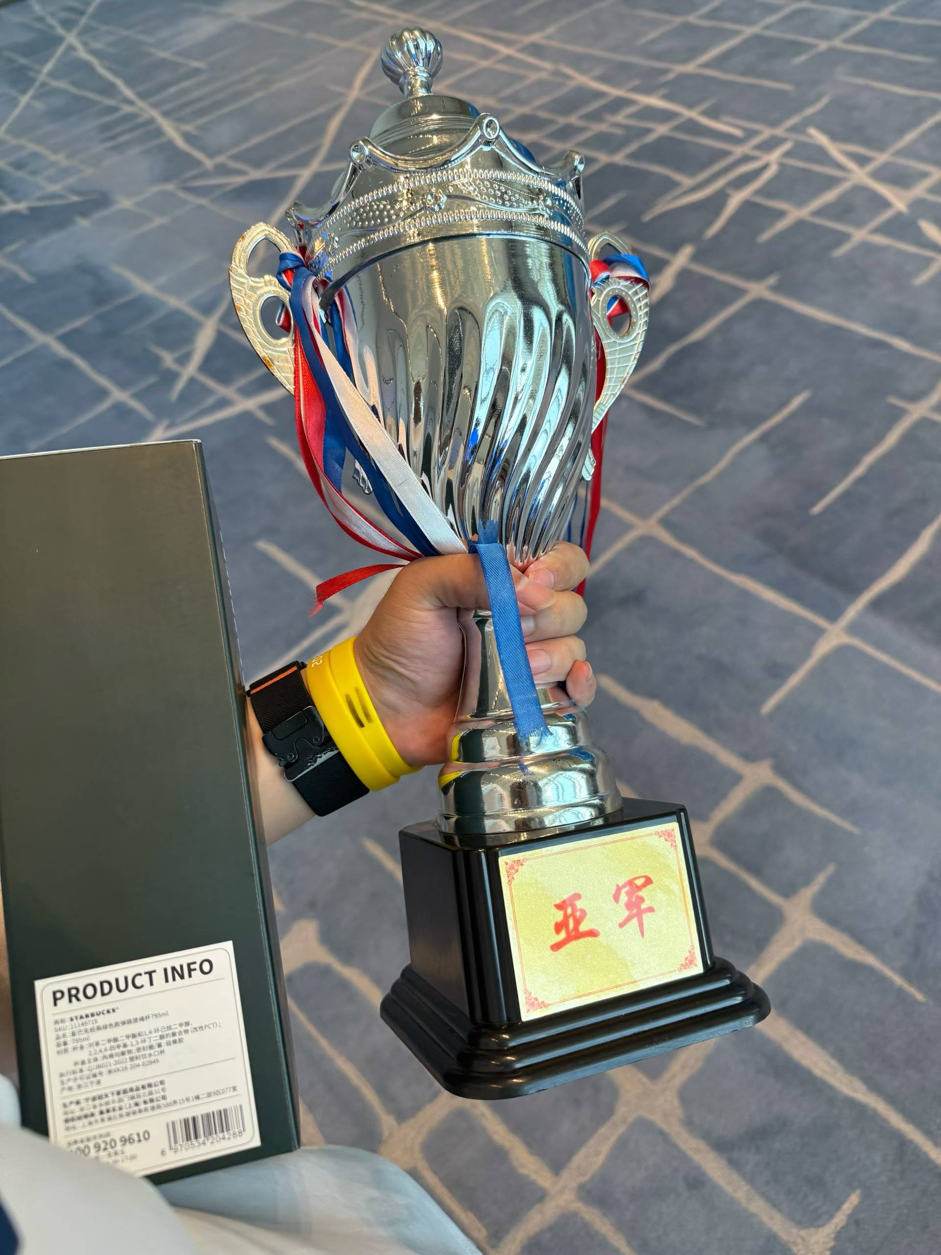 A hand holding a silver runner-up trophy decorated with red, white, and blue ribbons.
